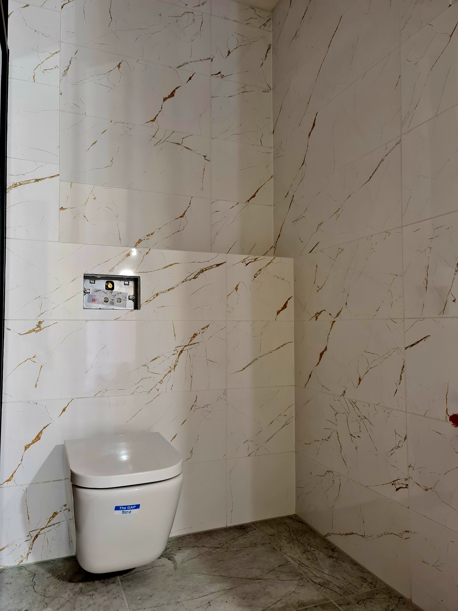 Świeżo wykafelkowana ściana łazienki z białymi płytkami imitującymi marmur z brązowymi żyłkami, widoczna podtynkowa instalacja sanitarna oraz podwieszana toaleta Roca The Gap.