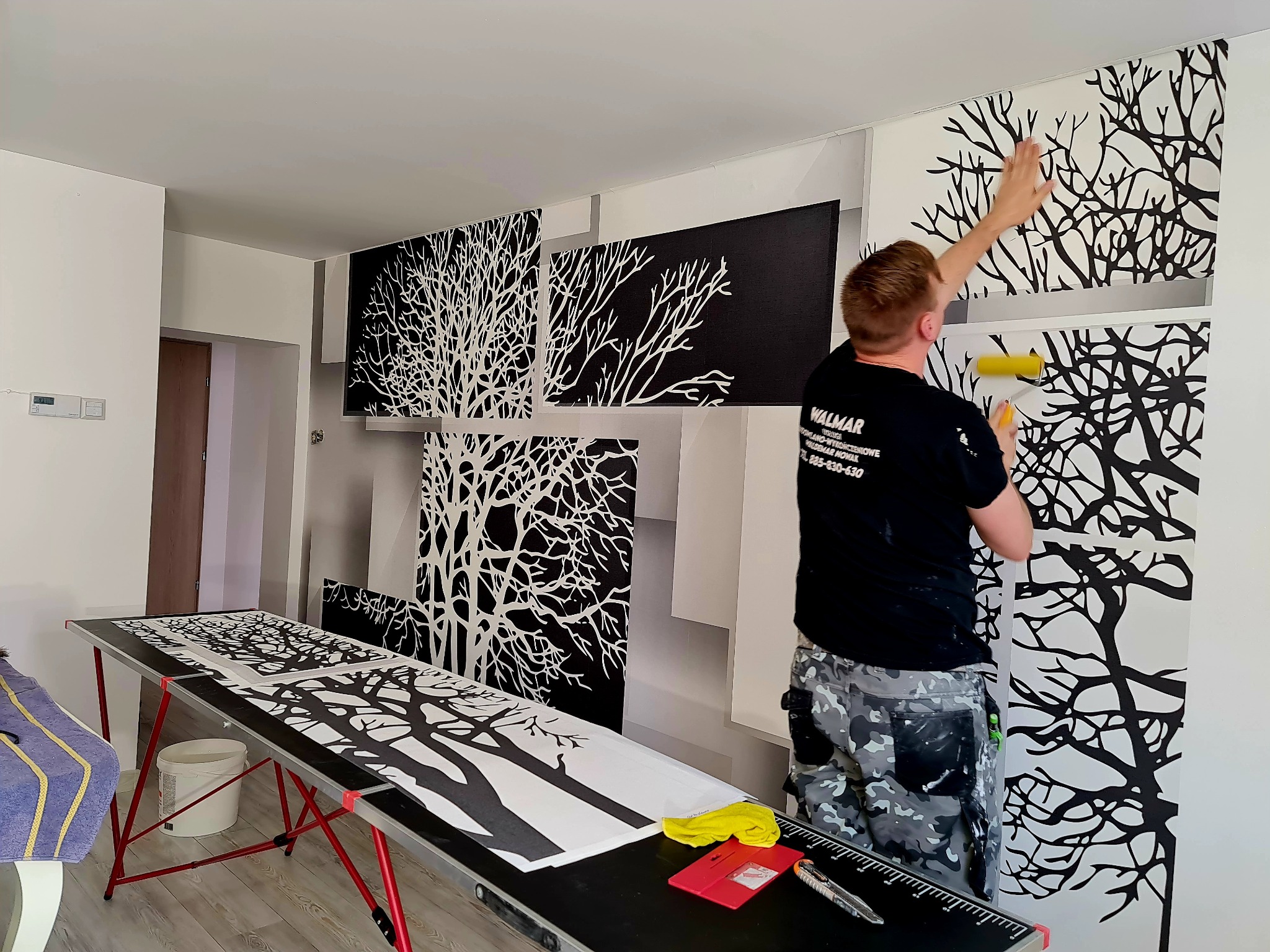 Pracownik w czarnej koszulce z logo firmy nakłada wałkiem tapetę z motywem drzewa na ścianę, widoczne panele tapety ułożone na stole, narzędzia pracy i wiadro z klejem.