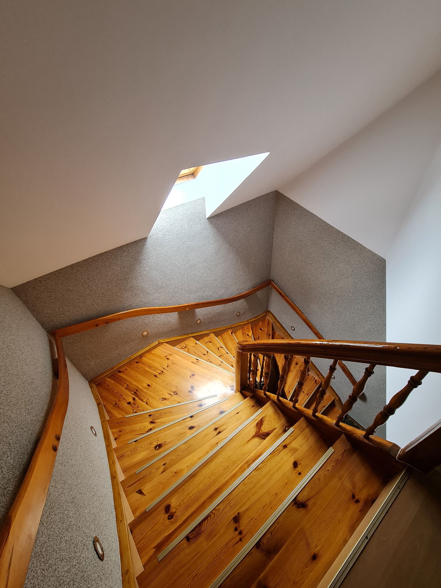 Drewniane spiralne schody z jasnymi stopniami i drewnianą balustradą, prowadzące do góry, oświetlone światłem wpadającym przez okno dachowe, ściany wykończone szarą tapetą strukturalną i punktowym...