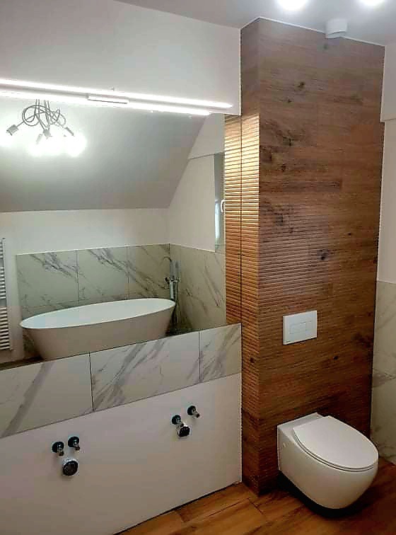 Nowoczesna łazienka na poddaszu z wolnostojącą wanną obudowaną płytami imitującymi marmur i toaletą podwieszaną na tle ściany wyłożonej panelami drewnopodobnymi z poziomymi żłobieniami.