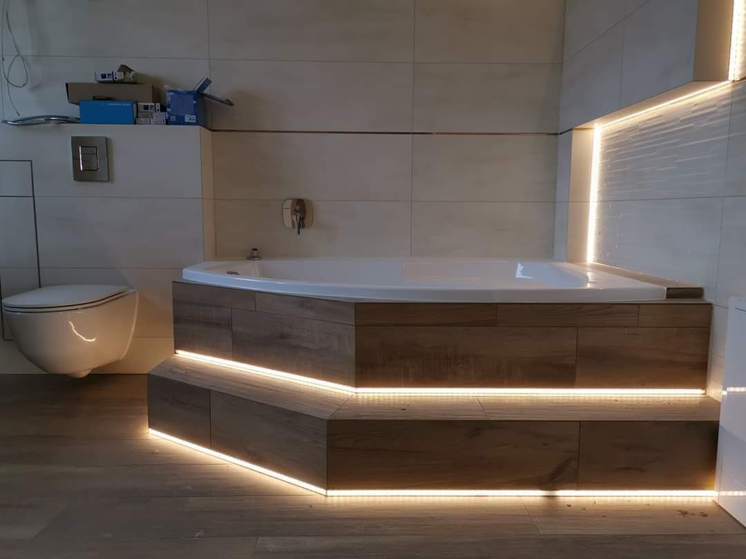 Nowoczesna łazienka z podświetlaną wanną narożną obudowaną drewnopodobnymi płytkami, wisząca toaleta w tle, oświetlenie LED wzdłuż wanny i w rogu pomieszczenia.