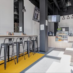 Centrum Konferencyjne Polska 13, Restauracja Odskocznia - Wnętrze nowoczesnej kawiarni biurowej z wysokim stołem barowym, czarnymi krzesłami, ekspozycją z napojami i przekąskami, oraz oświetleniem punktowym.