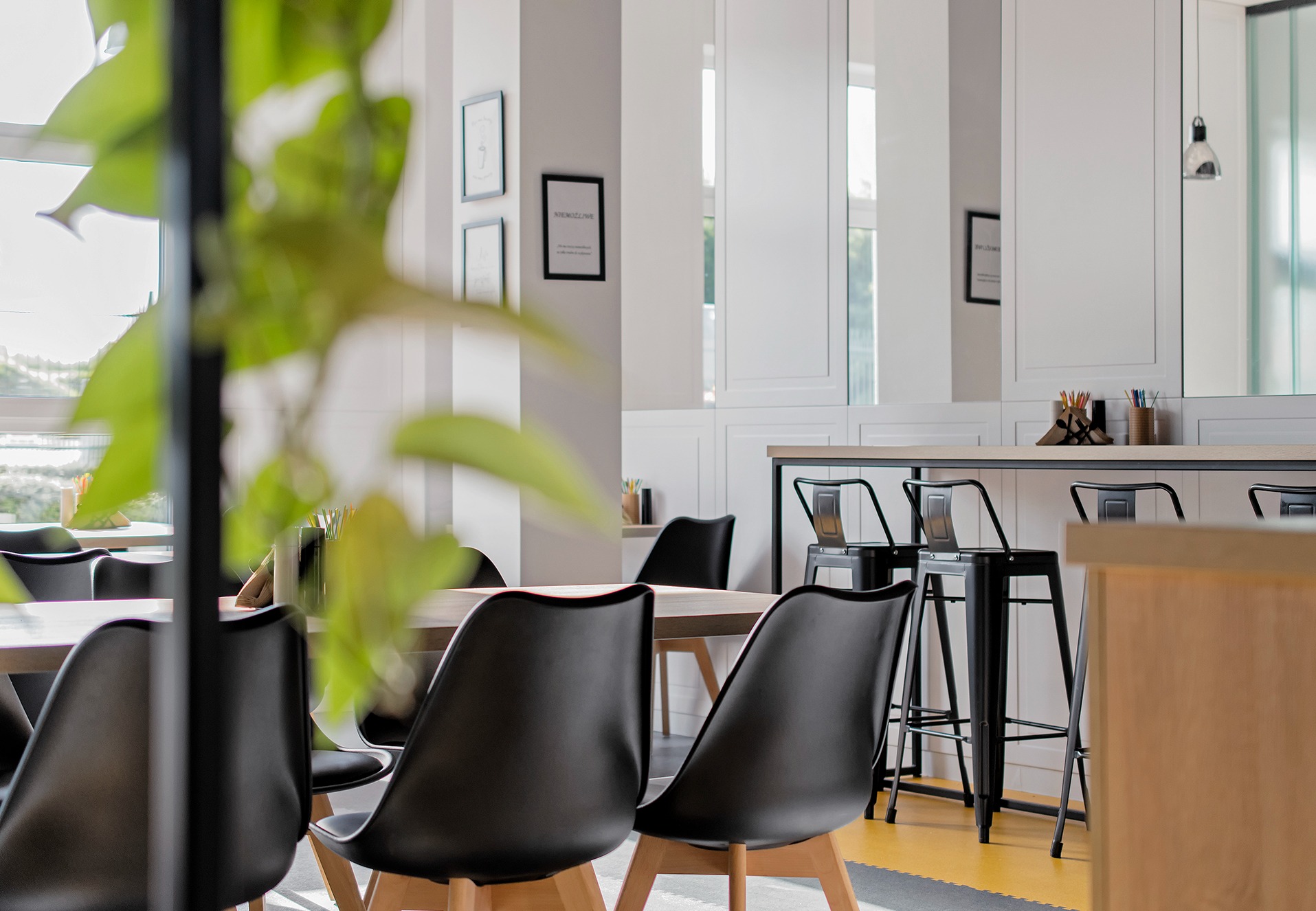 Jasne, nowoczesne wnętrze biurowe z drewnianym stołem i czarnymi krzesłami, widok przez roślinę doniczkową, w tle wysokie krzesła barowe i białe szafki.