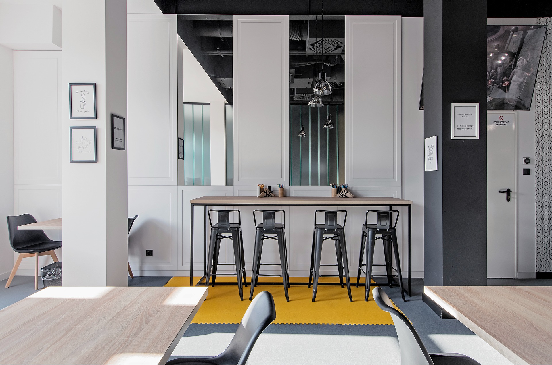 Nowoczesna aranżacja przestrzeni biurowej z wysokimi stołami barowymi, czarnymi stołkami i żółtą podłogą. Na ścianie grafiki i napisy motywacyjne.