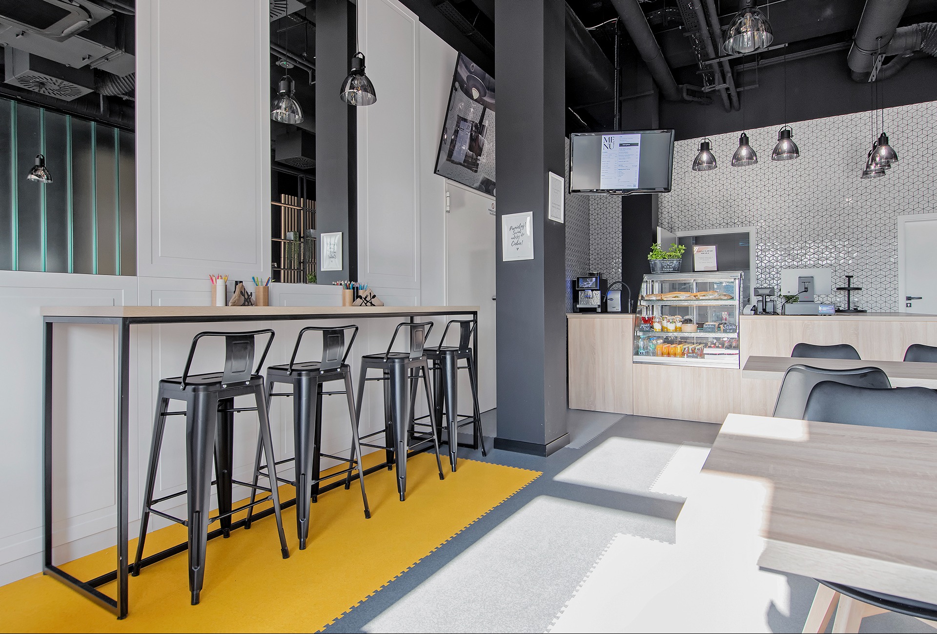 Wnętrze nowoczesnej kawiarni biurowej z wysokim stołem barowym, czarnymi krzesłami, ekspozycją z napojami i przekąskami, oraz oświetleniem punktowym.