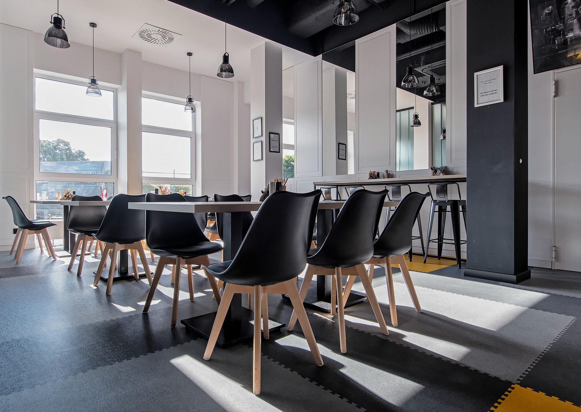 Jasne wnętrze restauracji lub sali konferencyjnej z czarnymi krzesłami i stołami, widoczne okna i oświetlenie sufitowe, podłoga z puzzli w odcieniach szarości i żółci.