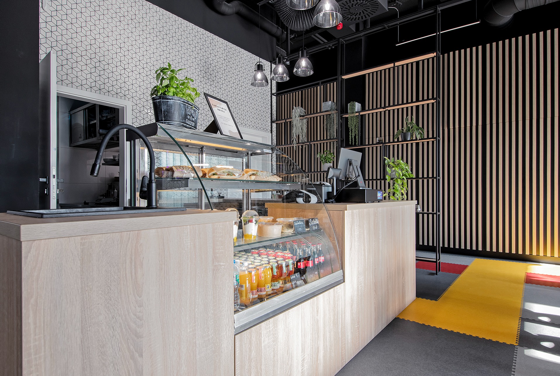 Nowoczesna lada barowa z chłodzoną witryną, prezentująca napoje i przekąski, z elementami dekoracyjnymi jak roślina w doniczce i czarne lampy wiszące, na tle ściany z drewnianych listewek.