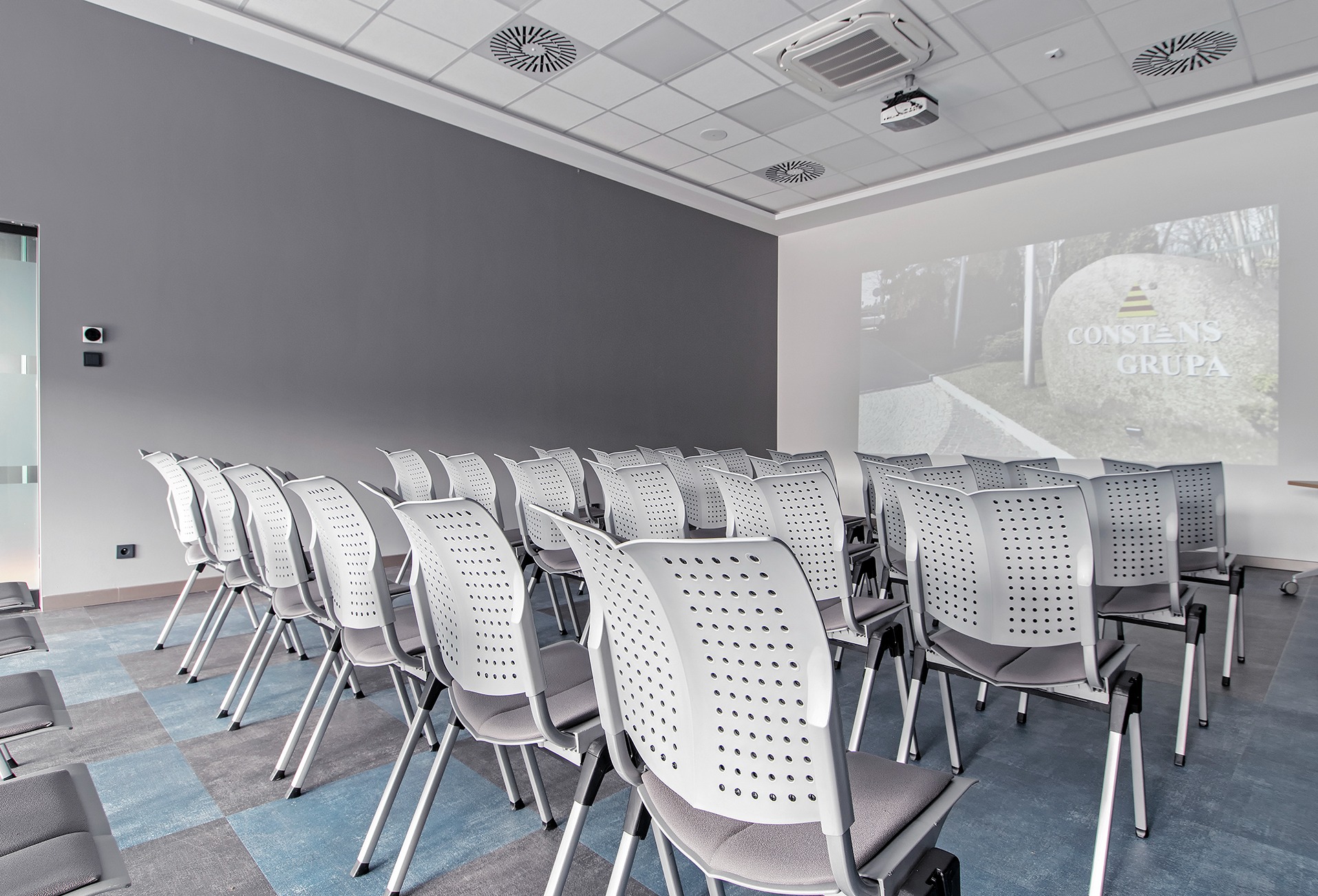 Sala konferencyjna z rzędami białych krzeseł z szarymi siedziskami, projektor wyświetlający logo firmy na ścianie, wentylacja sufitowa i klimatyzacja.