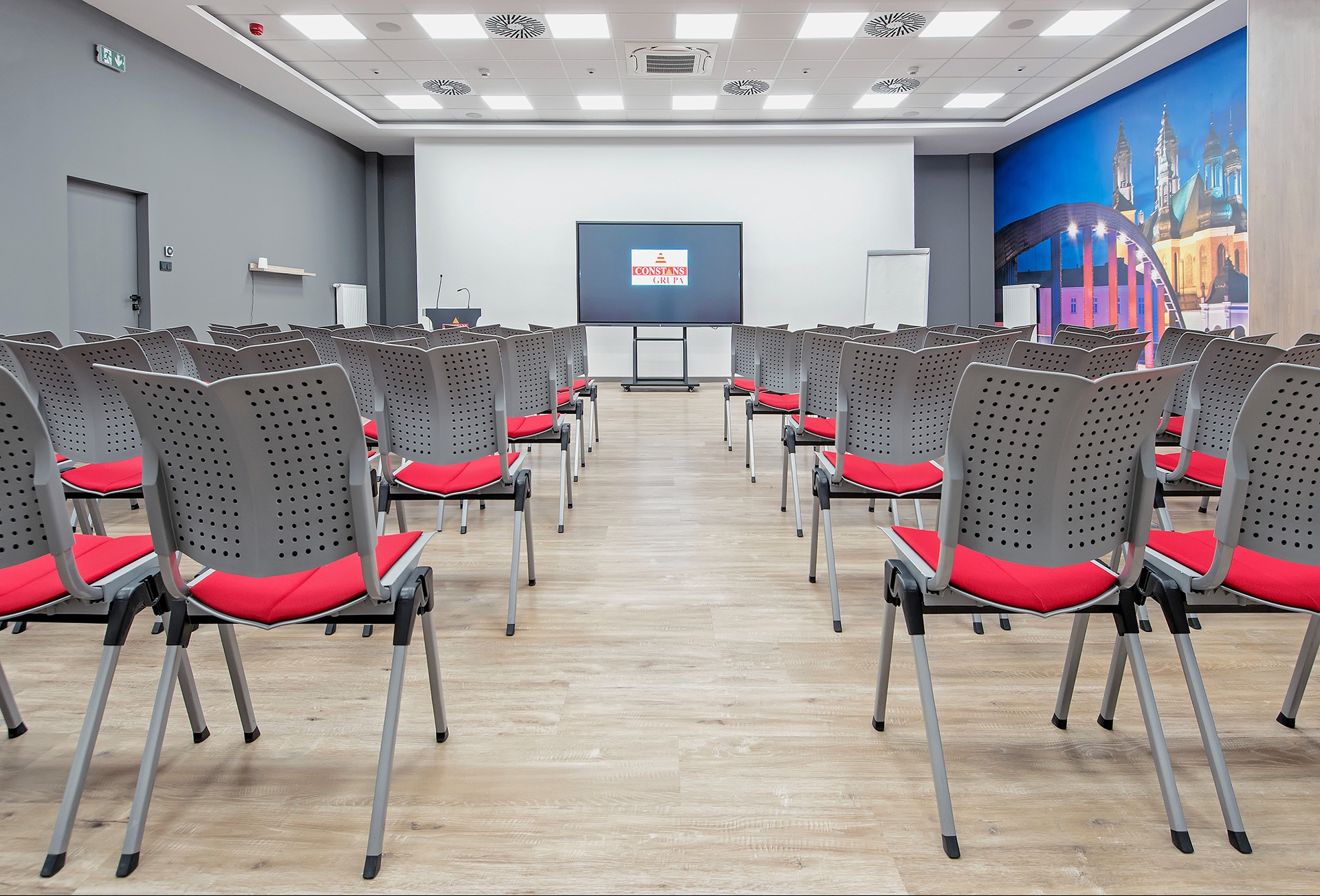 Sala konferencyjna z rzędami szarych krzeseł z czerwonymi siedziskami, ekranem wyświetlającym logo firmy i muralem z panoramą miasta na ścianie.