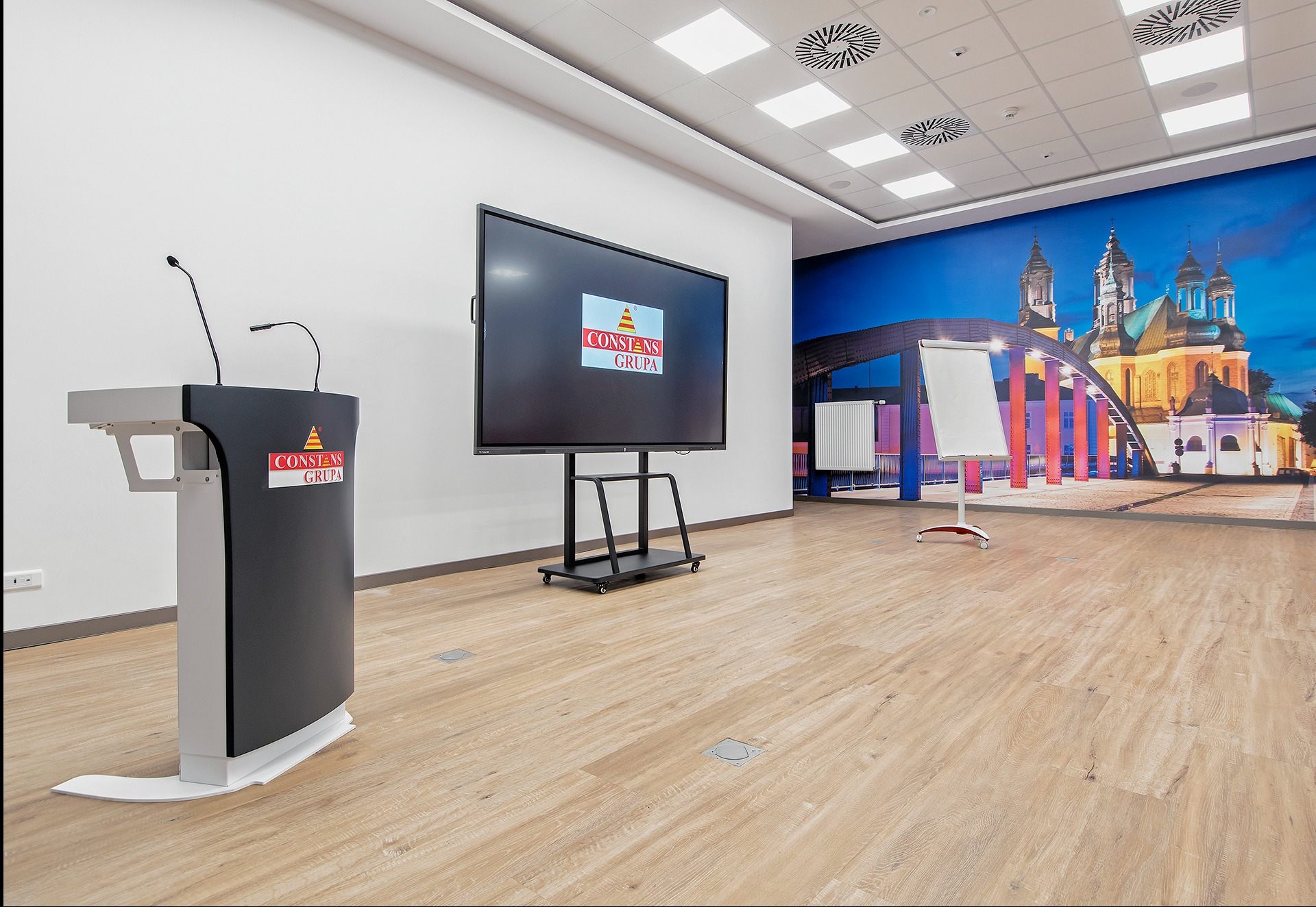 Sala konferencyjna z mównicą z logo Constans Grupa, ekranem wyświetlającym logo firmy i tłem z panoramą miasta.