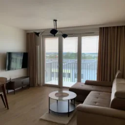 Jasne wnętrze salonu z brązową sofą narożną, stolikiem kawowym z okrągłym szklanym blatem na dywanie, stołem z krzesłami w tle oraz dużymi oknami balkonowymi z żaluzjami i brązowymi zasłonami.