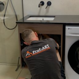 Prof-ART - Hydraulik w koszulce firmowej 'Prof-ART' naprawia instalację pod umywalką z czarnym blatem w łazience obok pralki LG.