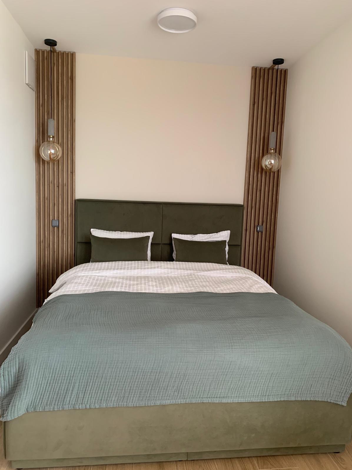 Nowoczesna sypialnia z łóżkiem w odcieniach zieleni, drewnianymi panelami dekoracyjnymi po bokach i wiszącymi lampami z żarówkami Edisona. Minimalistyczny design z okrągłą lampą sufitową.