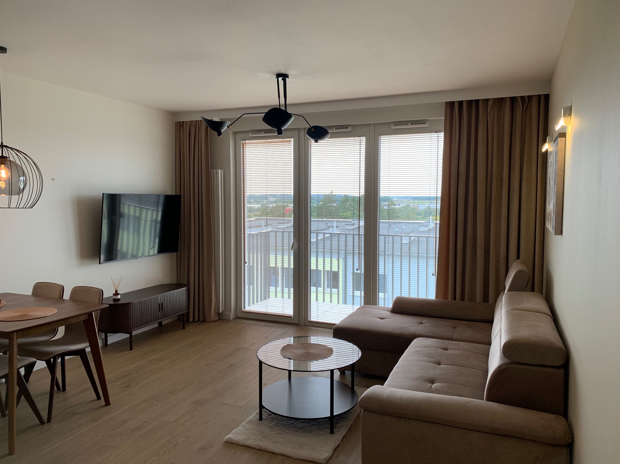 Jasne wnętrze salonu z brązową sofą narożną, stolikiem kawowym z okrągłym szklanym blatem na dywanie, stołem z krzesłami w tle oraz dużymi oknami balkonowymi z żaluzjami i brązowymi zasłonami.