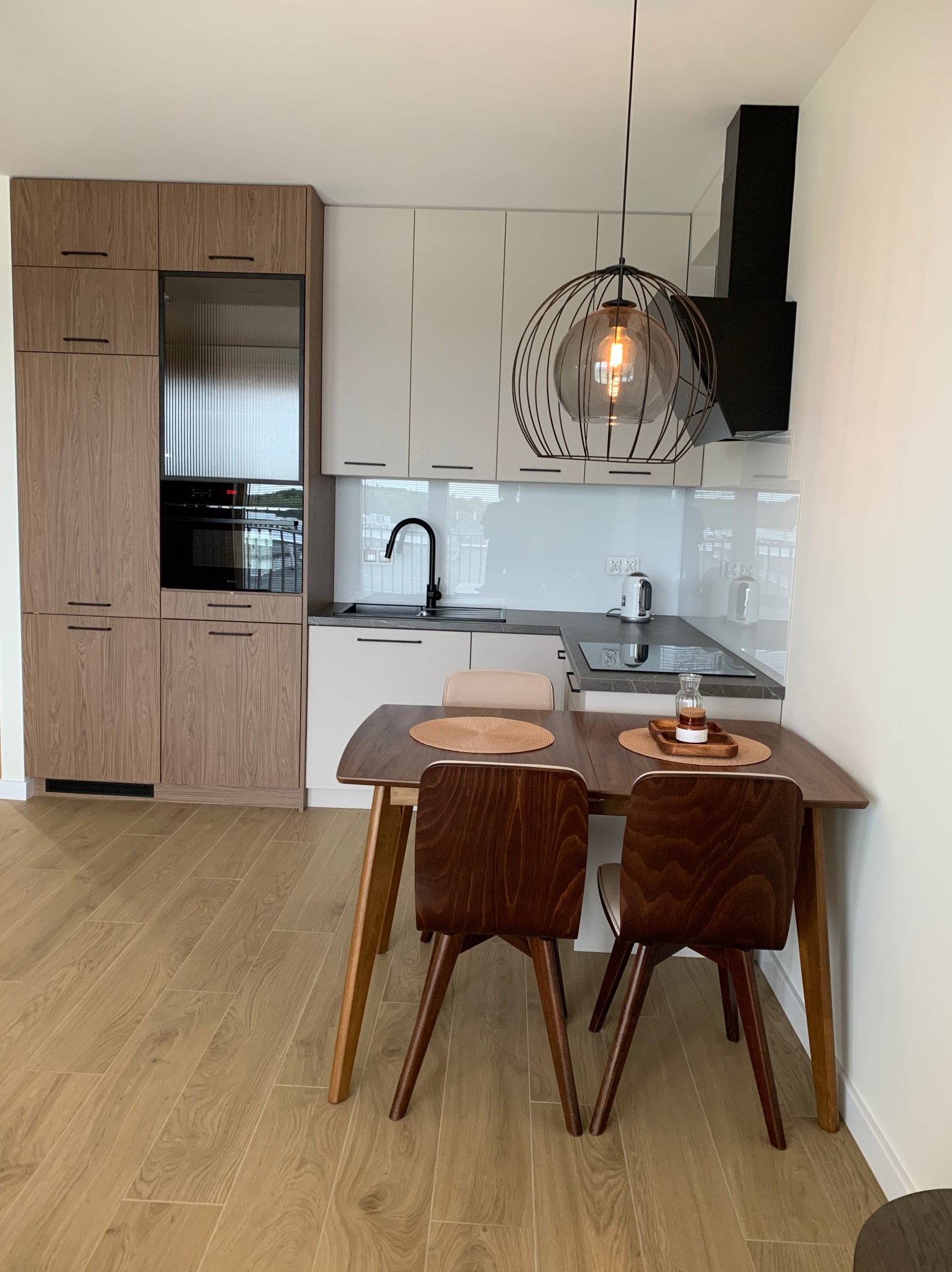 Nowoczesna kuchnia z drewnianymi i białymi szafkami, czarną armaturą i okapem, oświetlona dekoracyjną lampą wiszącą nad drewnianym stołem z brązowymi krzesłami, na podłodze panele imitujące drewno.