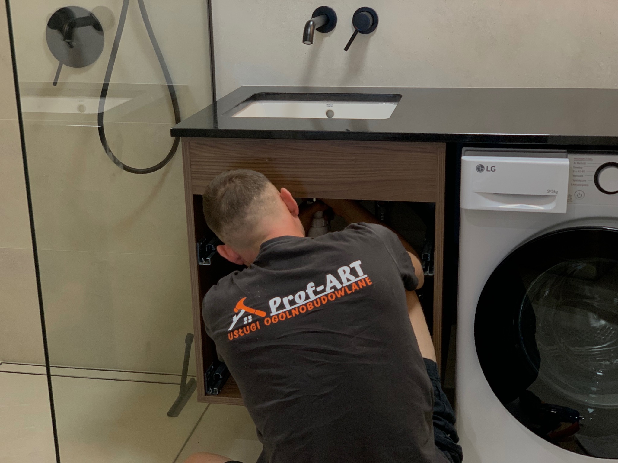 Hydraulik w koszulce firmowej 'Prof-ART' naprawia instalację pod umywalką z czarnym blatem w łazience obok pralki LG.