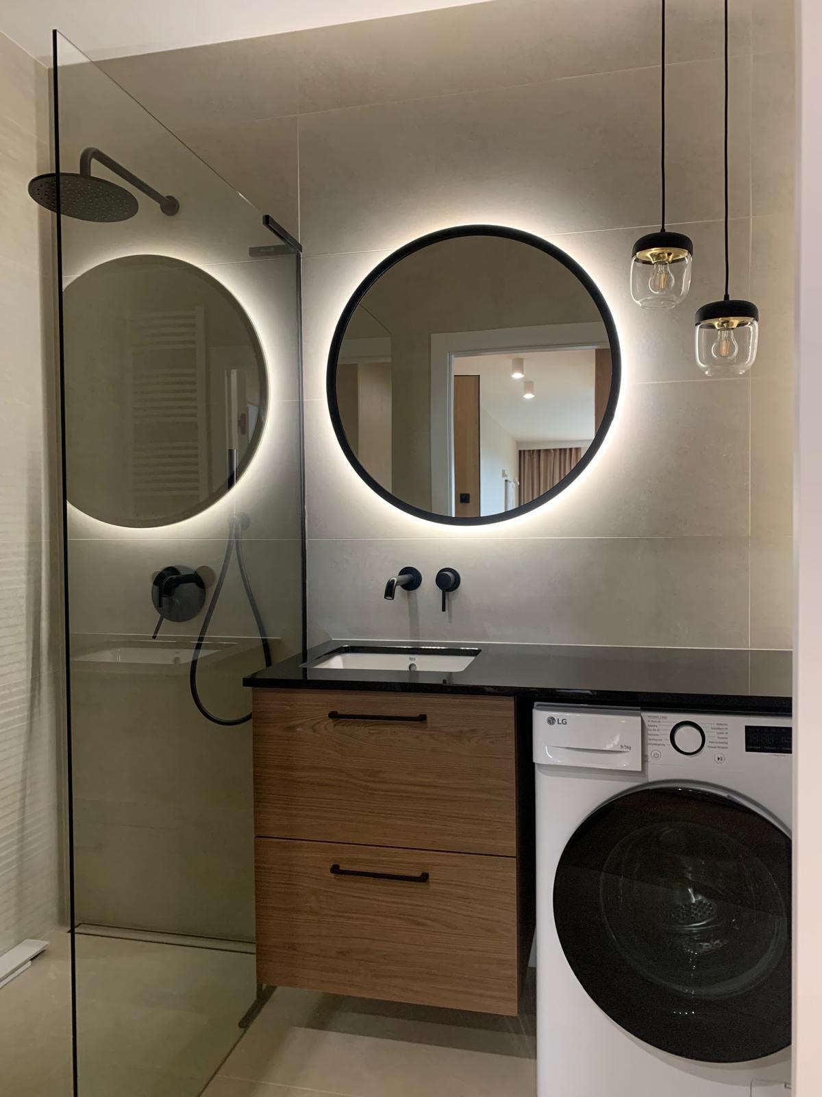 Nowoczesna łazienka z okrągłymi lustrami podświetlanymi LED, czarną armaturą, szafką pod umywalką z drewnianym frontem i pralką LG.