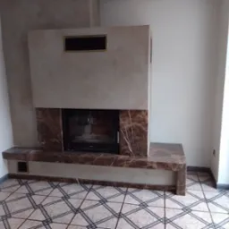 Wykończony kominek z marmurowym obramowaniem i ozdobną wnęką, umieszczony w rogu pomieszczenia z wzorzystą podłogą, obok okna balkonowego. Modernistyczny design.