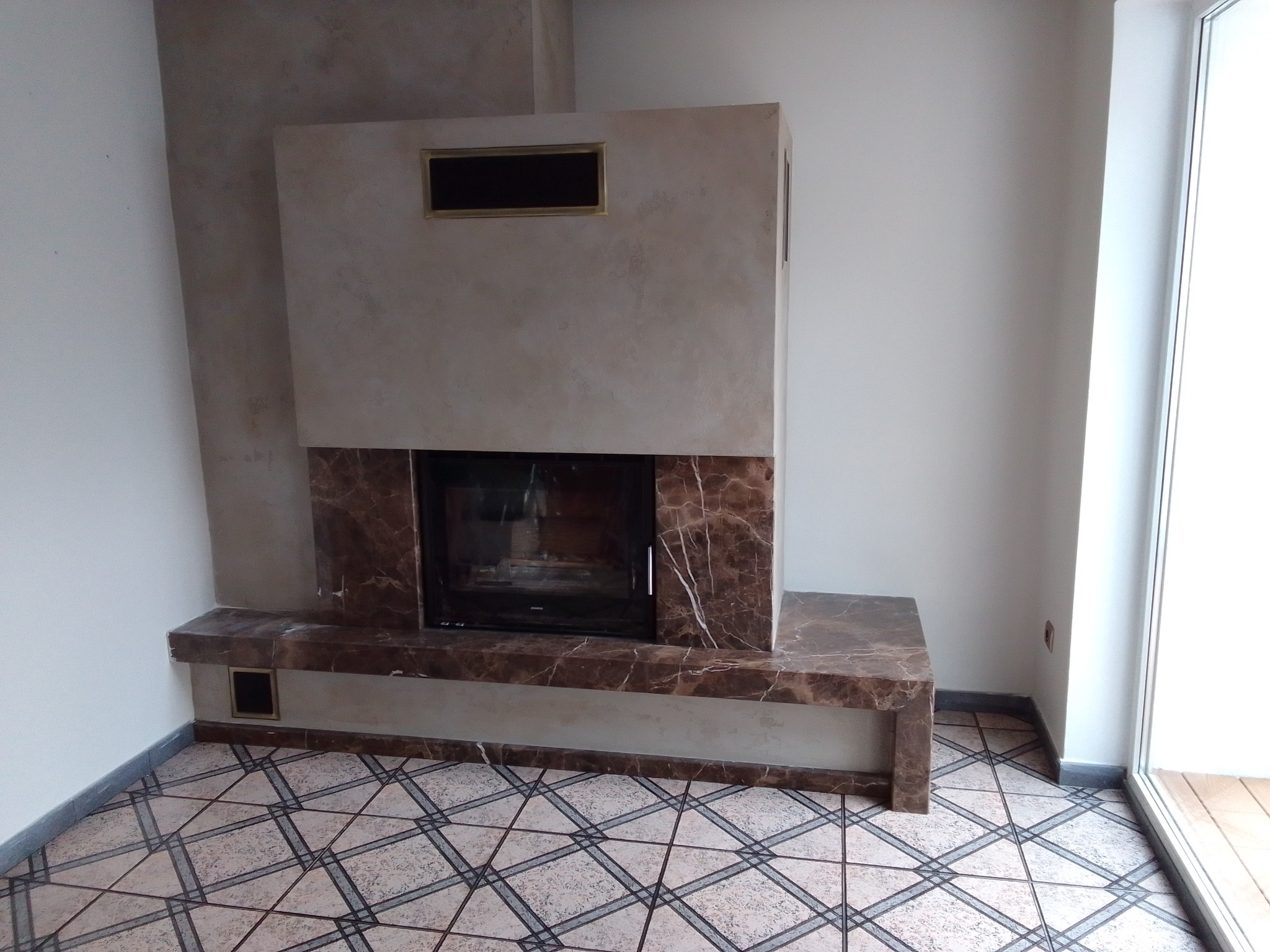 Wykończony kominek z marmurowym obramowaniem i ozdobną wnęką, umieszczony w rogu pomieszczenia z wzorzystą podłogą, obok okna balkonowego. Modernistyczny design.