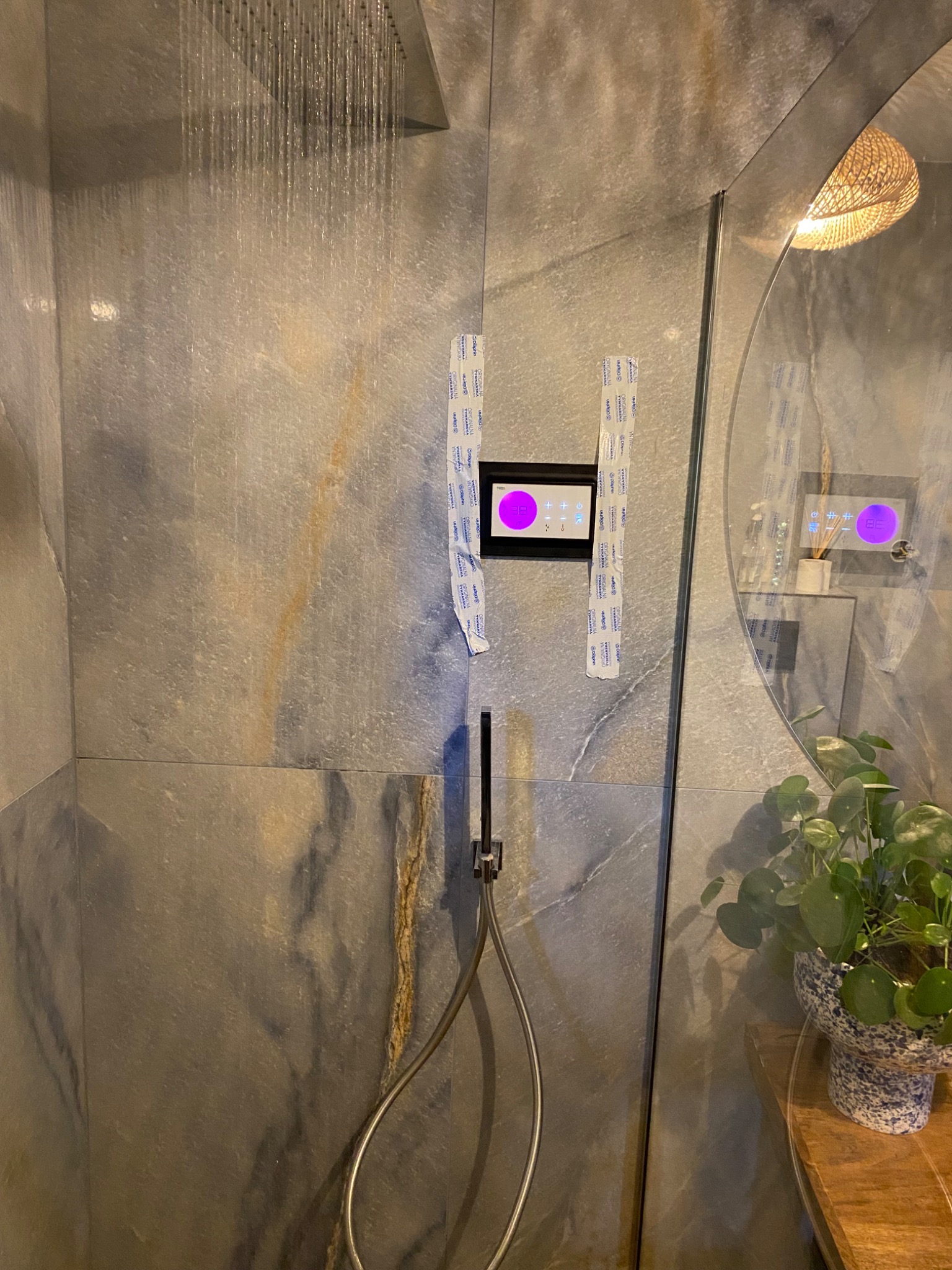 Nowoczesna kabina prysznicowa z deszczownicą, szare płytki imitujące kamień, dotykowy panel sterowania temperaturą zabezpieczony folią ochronną, okrągłe lustro i roślina doniczkowa na drewnianym...