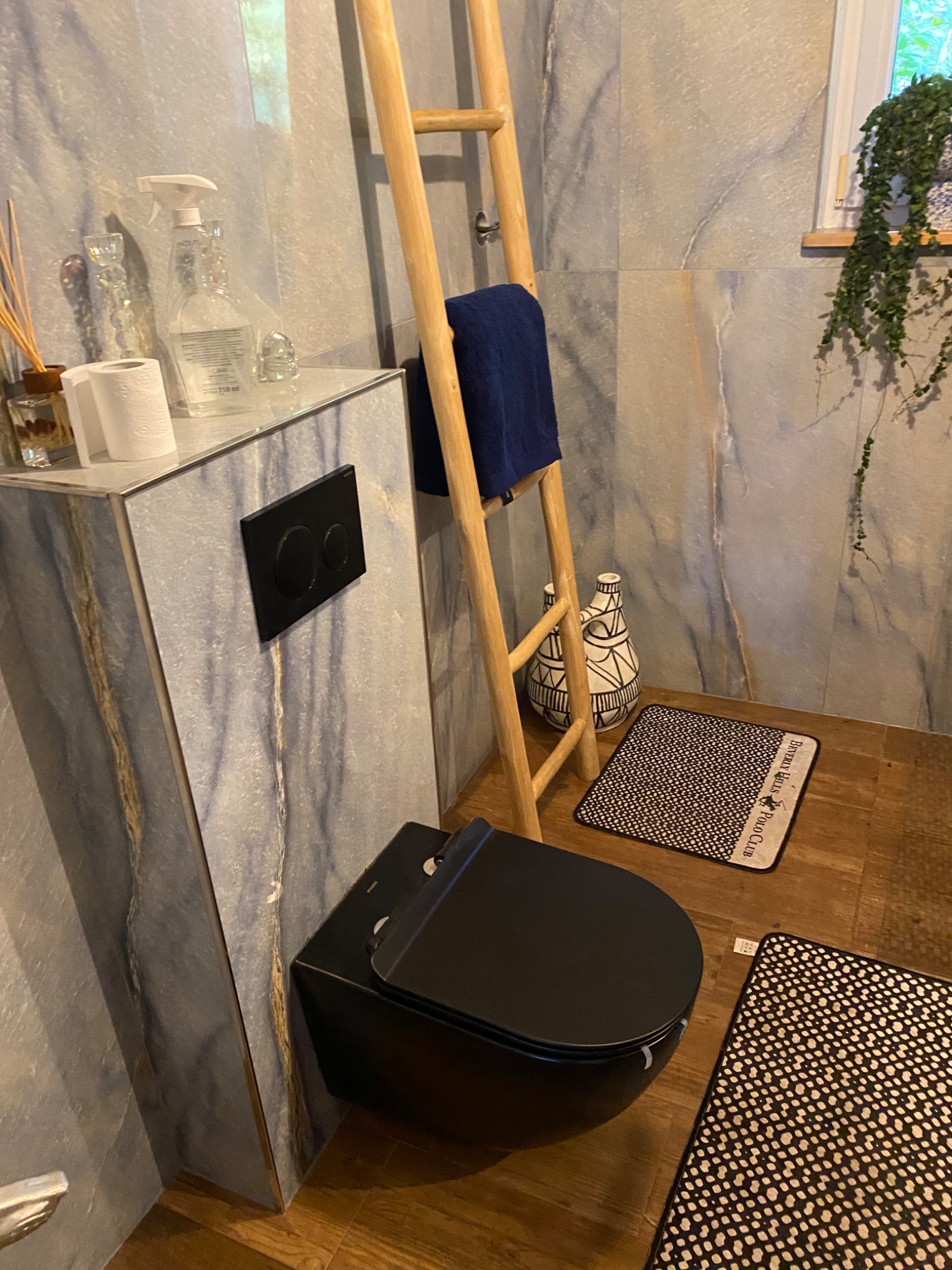Nowoczesna łazienka z wiszącą toaletą w kolorze czarnym, marmurowe płytki na ścianach, drewniana drabina z niebieskim ręcznikiem, dywaniki w czarno-białe wzory i dekoracyjna doniczka z rośliną.