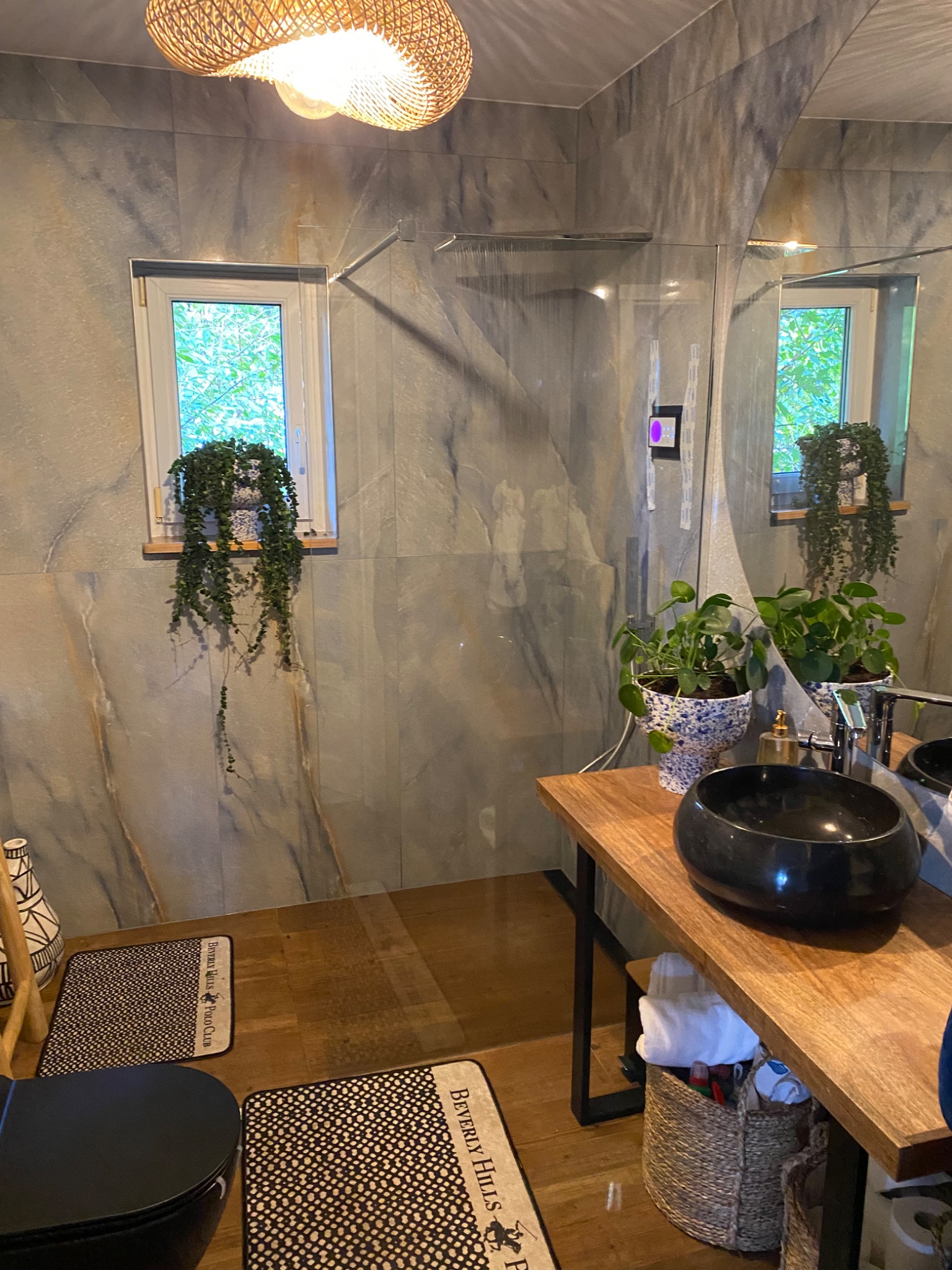 Nowoczesna łazienka z marmurowymi płytkami, wiszącą rośliną przy oknie, czarną umywalką na drewnianym blacie i plecioną lampą sufitową.