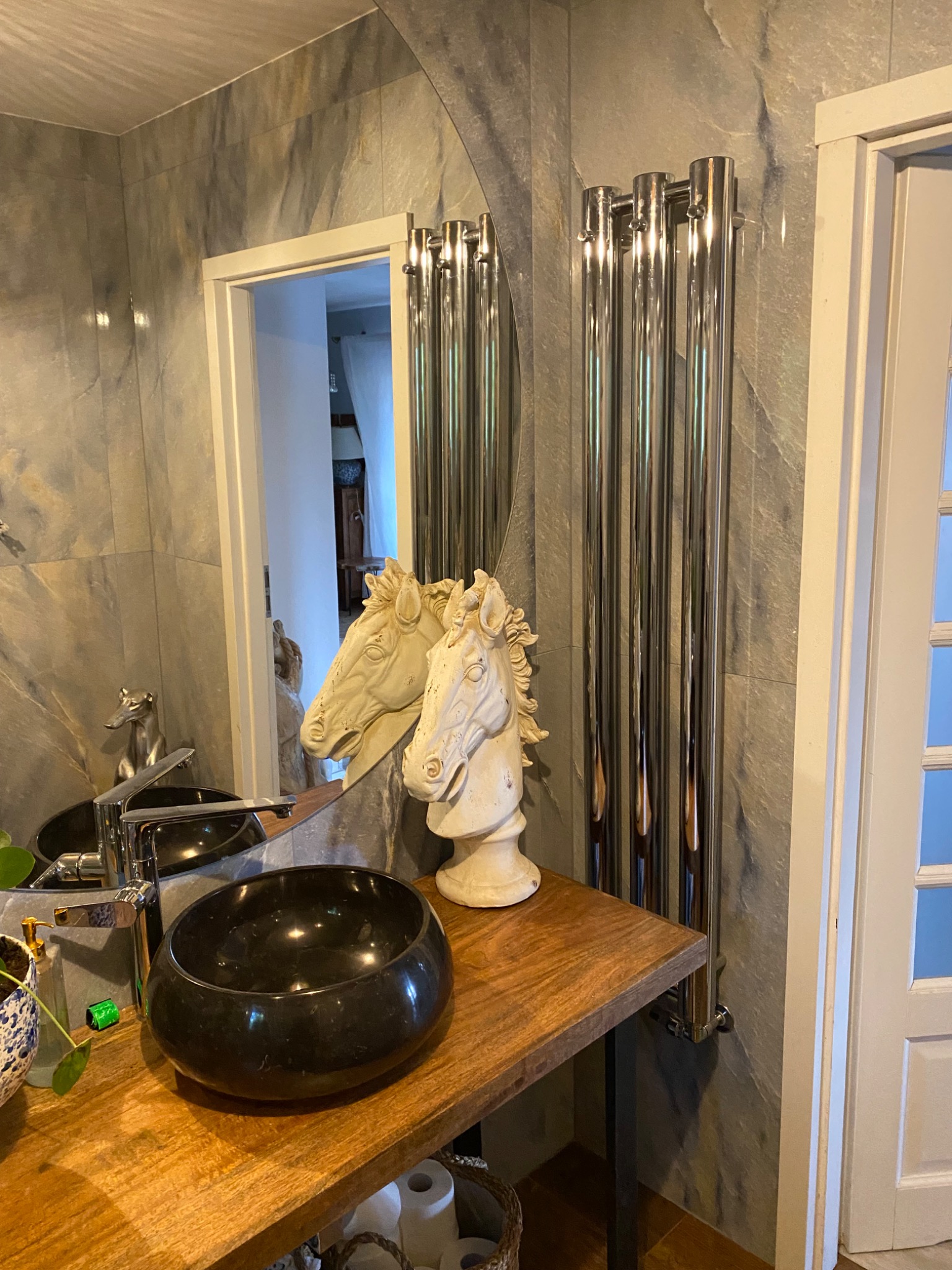 Czarna umywalka nablatowa o owalnym kształcie na drewnianym blacie w łazience z szarymi płytkami imitującymi kamień, obok chromowany grzejnik drabinkowy i dekoracyjne figurki końskich głów.