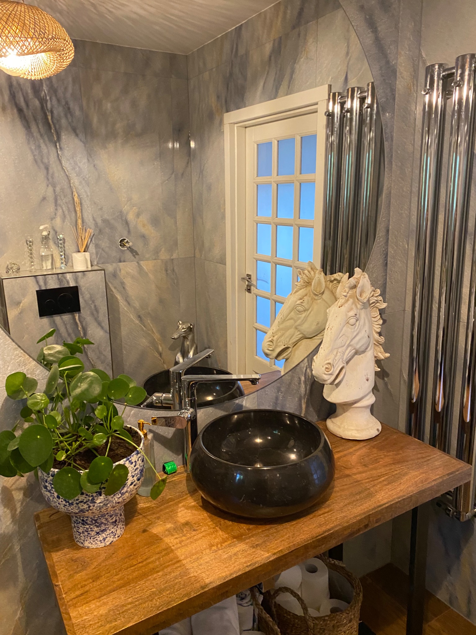 Nowoczesna łazienka z marmurowymi płytkami, okrągłym lustrem, drewnianym blatem z czarną umywalką, dekoracyjną figurką głowy konia i rośliną w niebieskiej doniczce.