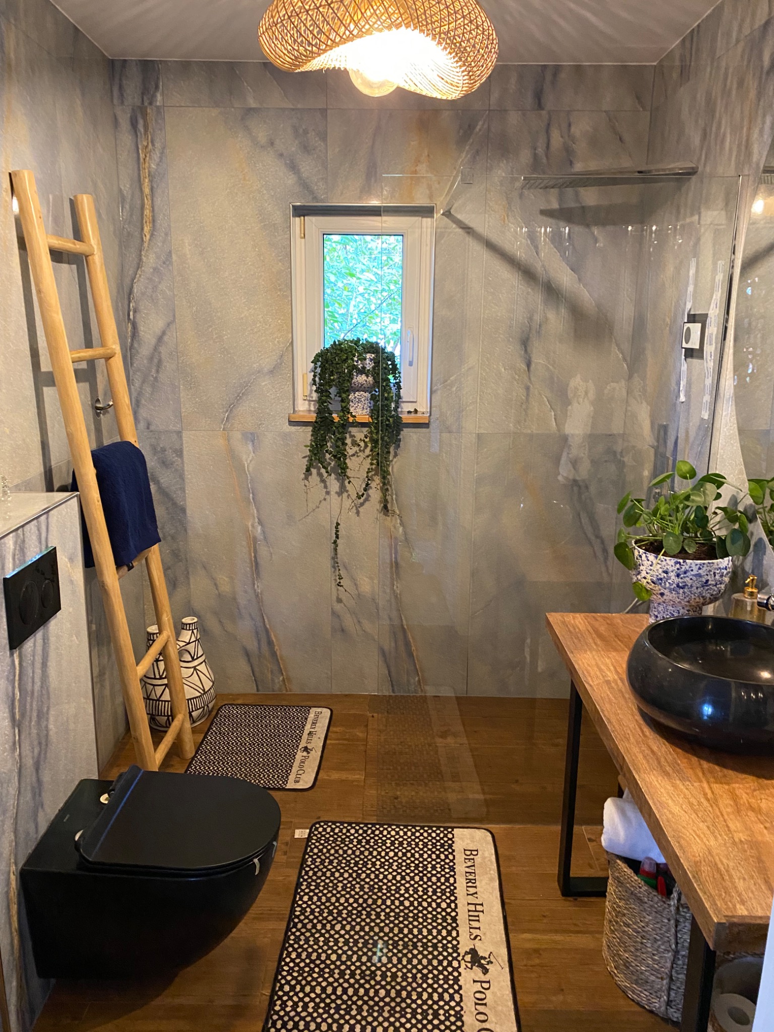 Nowoczesna łazienka z szarymi płytkami imitującymi marmur, drewnianą drabiną jako wieszak na ręczniki, wiszącą toaletą w kolorze czarnym oraz drewnianym blatem z czarną umywalką, oświetlona...