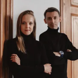 Portret kobiety i mężczyzny w czarnych golfach, stojących na tle drewnianych drzwi z dekoracyjnymi listwami i białej ściany.