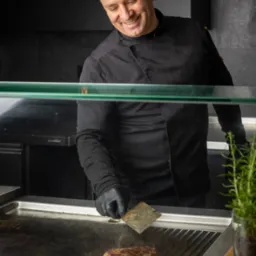 Uśmiechnięty kucharz w czarnym uniformie i rękawiczkach smaży stek na grillu, widok zza szyby ekspozycyjnej.