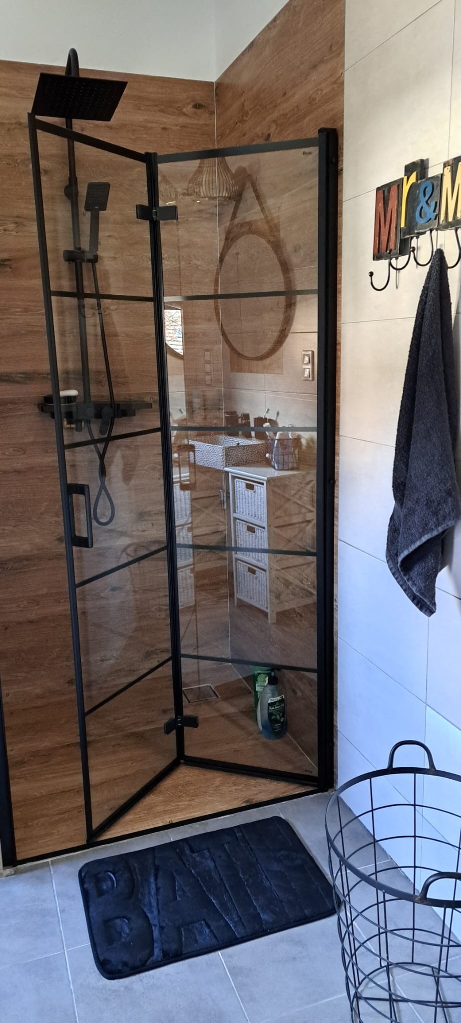 Nowoczesna kabina prysznicowa z czarnymi profilami, drewnopodobne płytki na ścianach, szary ręcznik na wieszaku i czarny kosz na pranie w tle.