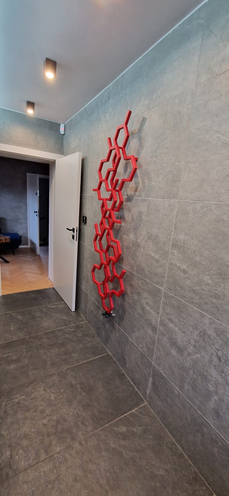 Szare płytki gresowe na ścianie i podłodze w korytarzu, z dekoracyjnym, czerwonym grzejnikiem w kształcie heksagonów. Widoczne drzwi wejściowe do pokoju z parkietem w jodełkę.