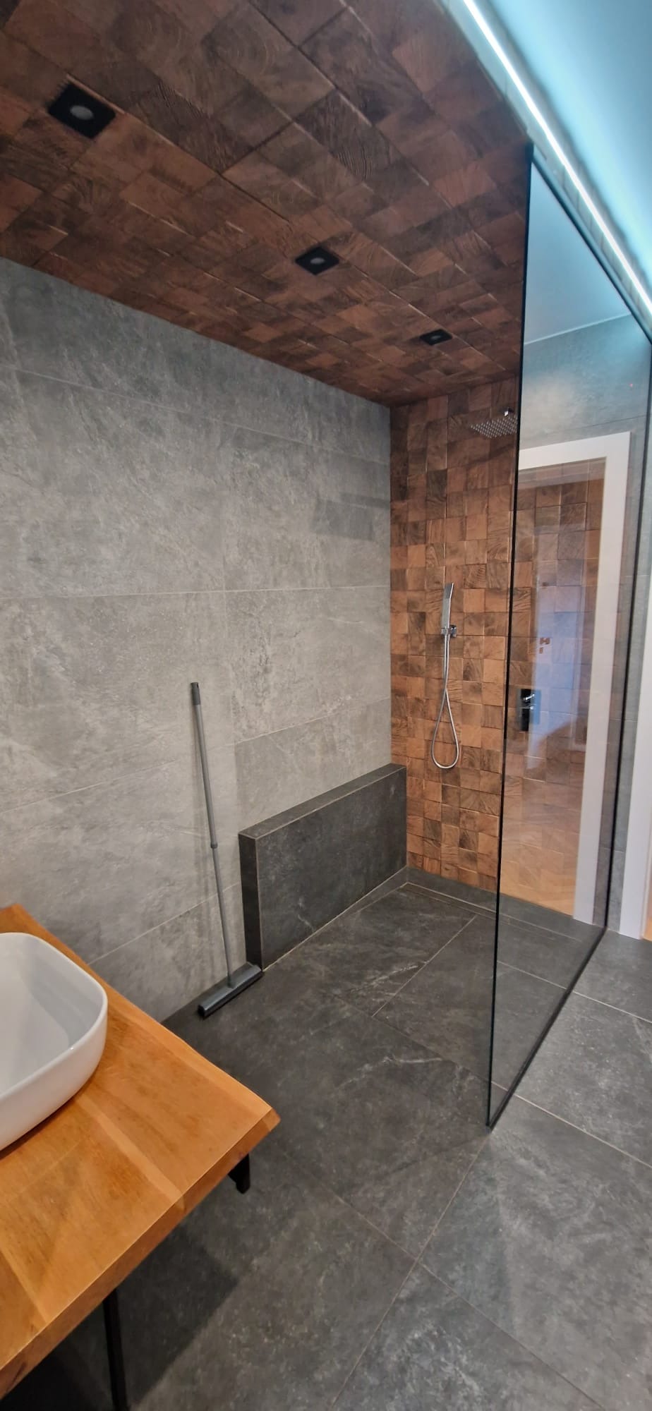 Nowoczesna łazienka z drewnianym sufitem, szarą kamienną ścianą i kabiną prysznicową typu walk-in z przezroczystego szkła. Widoczna drewniana umywalka i mop oparty o ścianę.