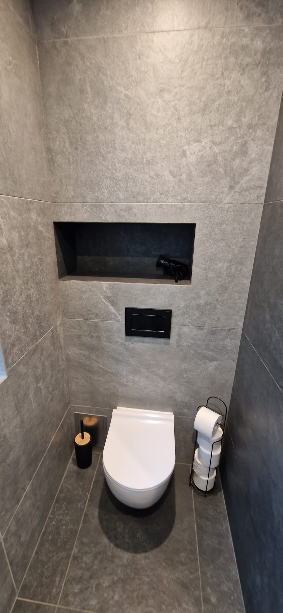 Nowoczesna toaleta z szarymi płytkami imitującymi kamień, podwieszana miska WC, czarny przycisk spłukujący i wnęka w ścianie z dekoracyjnym elementem.