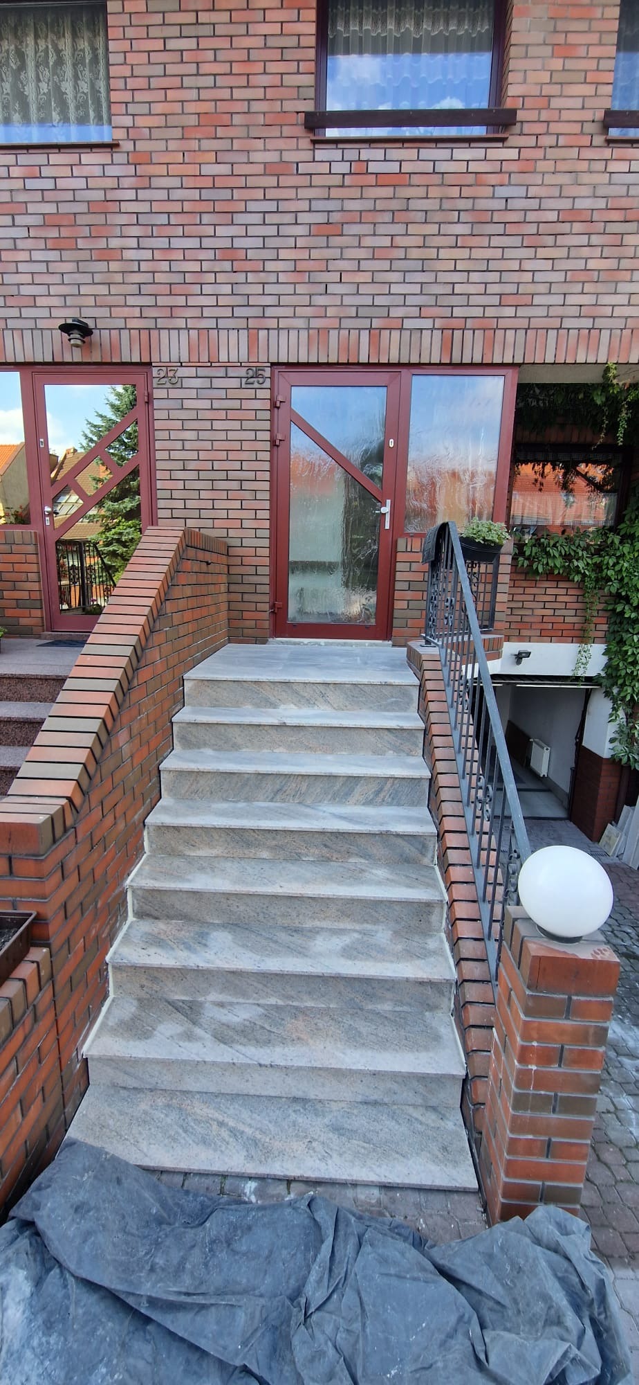 Zewnętrzne schody granitowe prowadzące do domu z czerwonej cegły, z metalową balustradą i kulistą lampą na słupku, częściowo zakryte folią ochronną.