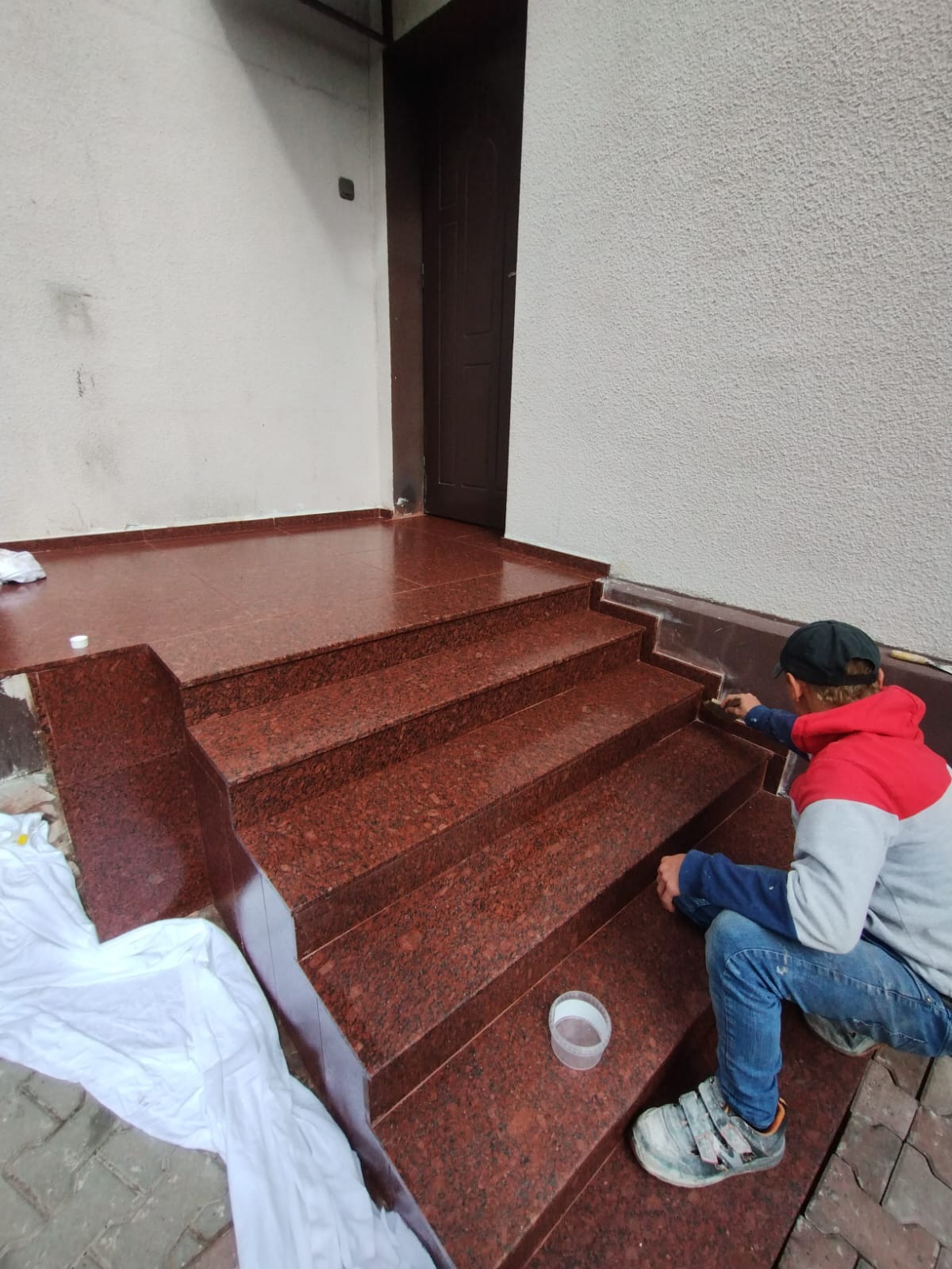 Montaż czerwonych schodów granitowych zewnętrznych, pracownik w bluzie z kapturem wykańcza spoiny przy ścianie, widoczne narzędzia i pojemnik na klej.