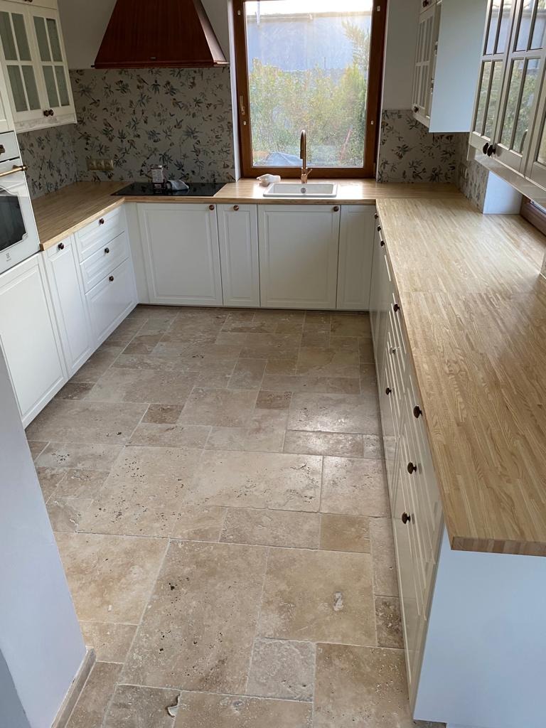 Kuchnia z posadzką trawertynową ułożoną z płytek o zróżnicowanej wielkości i naturalnym odcieniu beżu, widoczna zabudowa kuchenna w stylu rustykalnym z białymi szafkami i drewnianymi blatami.