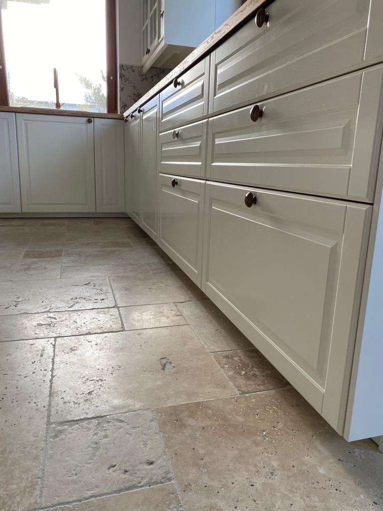 Beżowa posadzka z trawertynu w kuchni z kremowymi szafkami i drewnianym blatem, widok z niskiego kąta.
