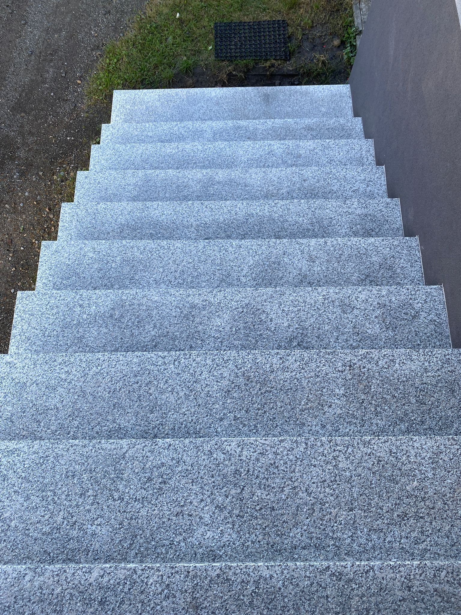 Zewnętrzne schody z szarego kamienia o szorstkiej fakturze, prowadzące w górę, widok z góry, obok trawnik i szara ściana.