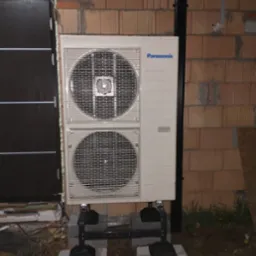 Zewnętrzna jednostka pompy ciepła Panasonic zamontowana na betonowych bloczkach i metalowej konstrukcji, widoczna na tle ściany z cegły dziurawki i czarnej rury spustowej, obok drzwi wejściowych.