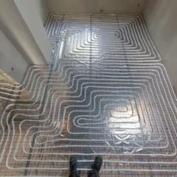 Rozłożone rury ogrzewania podłogowego na srebrnej folii izolacyjnej z czerwoną siatką, widok z góry na instalację w surowym pomieszczeniu z butami osoby wykonującej prace.