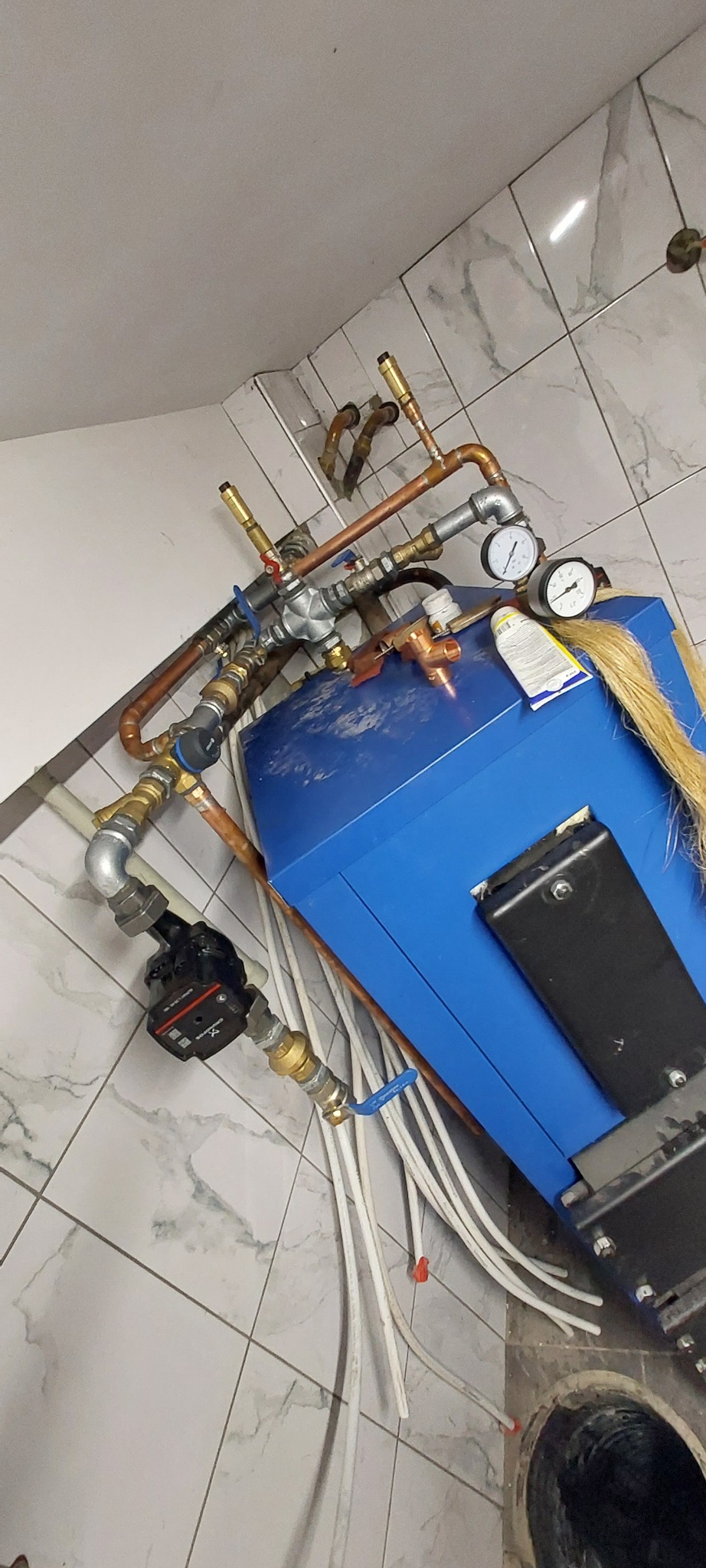 Niebieski zbiornik z podłączonymi rurami miedzianymi, manometrami i pompą obiegową, widoczne białe rury oraz fragment podłogi z otworem.