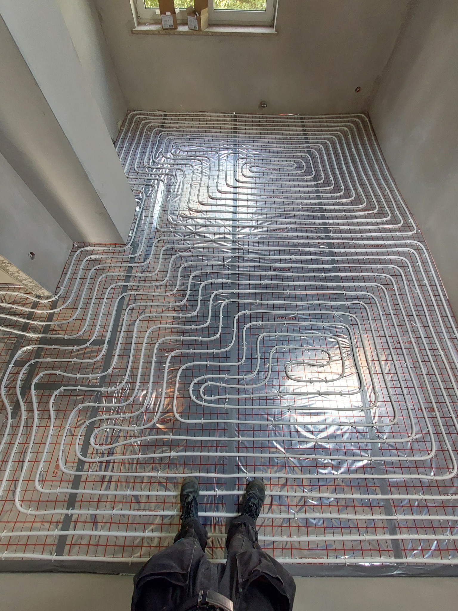 Rozłożone rury ogrzewania podłogowego na srebrnej folii izolacyjnej z czerwoną siatką, widok z góry na instalację w surowym pomieszczeniu z butami osoby wykonującej prace.