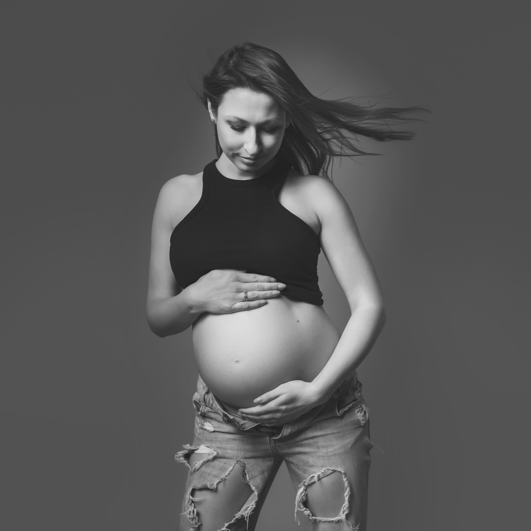 Czarno-białe zdjęcie kobiety w ciąży, z rozwianymi włosami, ubrana w czarny top i podarte jeansy, trzymająca ręce na brzuchu, patrząca w dół.