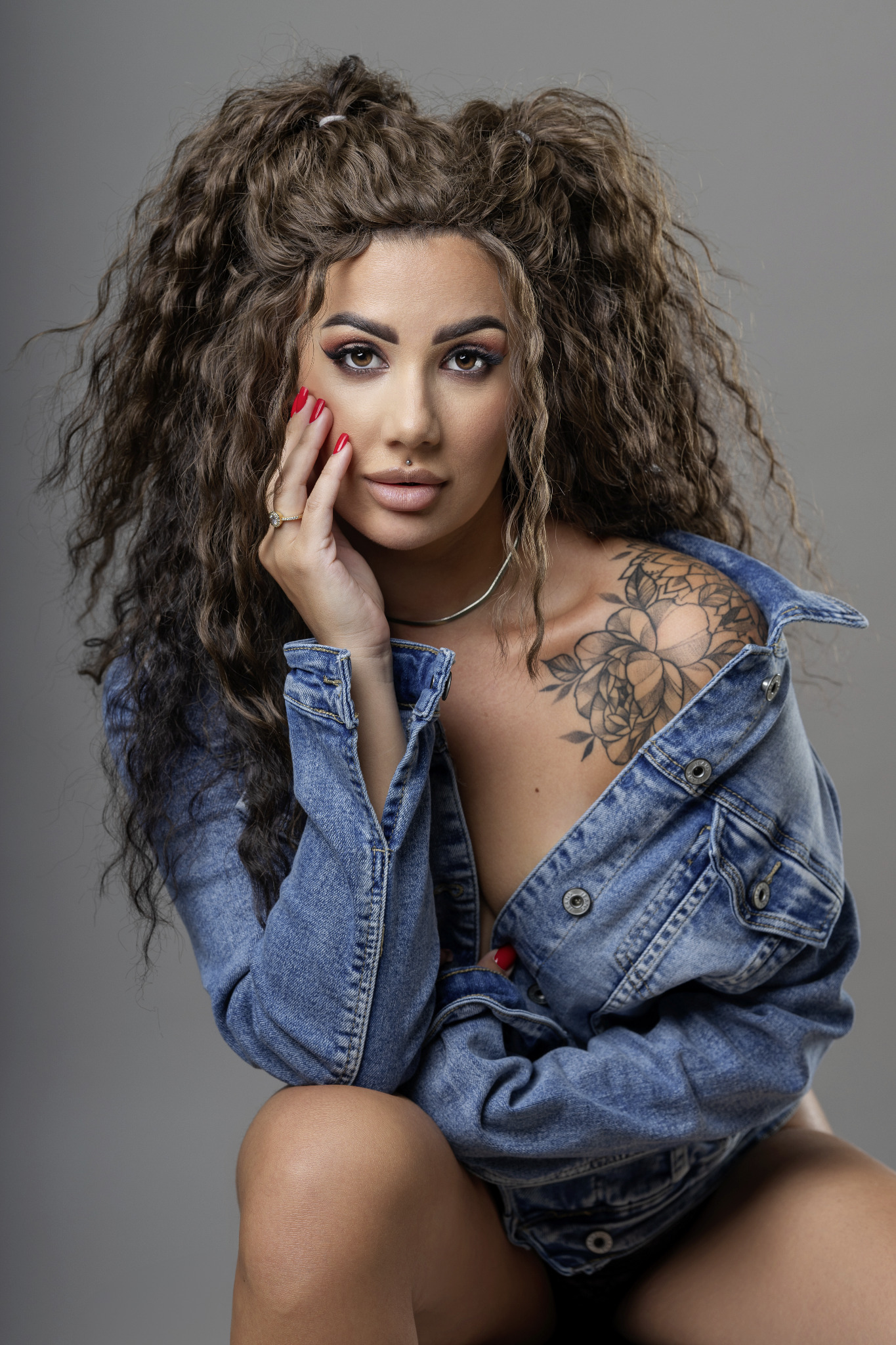 Portret kobiety z tatuażem na ramieniu, w rozpiętej kurtce jeansowej, z kręconymi włosami i kolczykiem w wardze, pozującej na szarym tle.