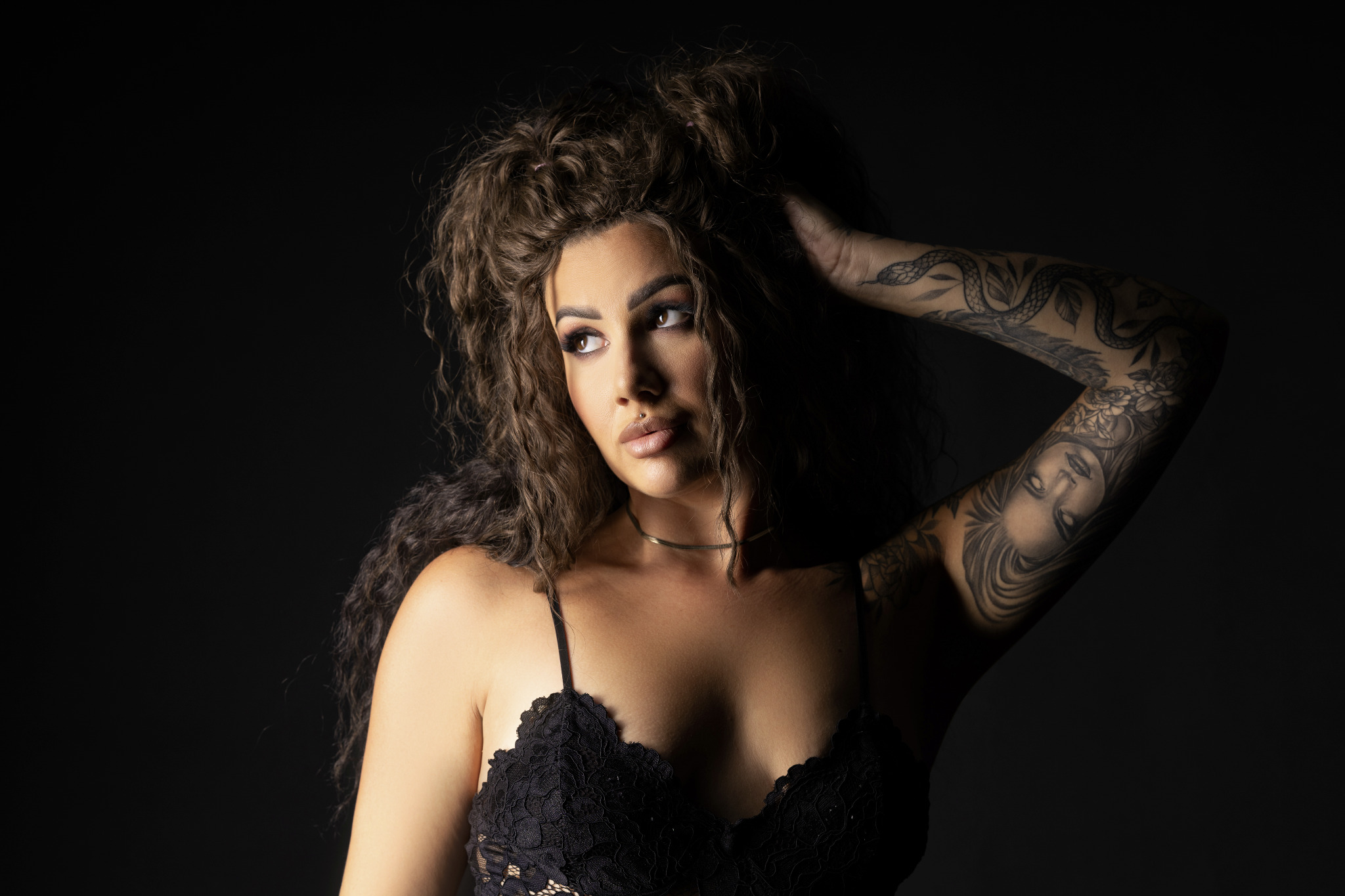 Portret kobiety z tatuażami na ramieniu, kręconymi włosami i piercingiem w wardze, pozującej na ciemnym tle.