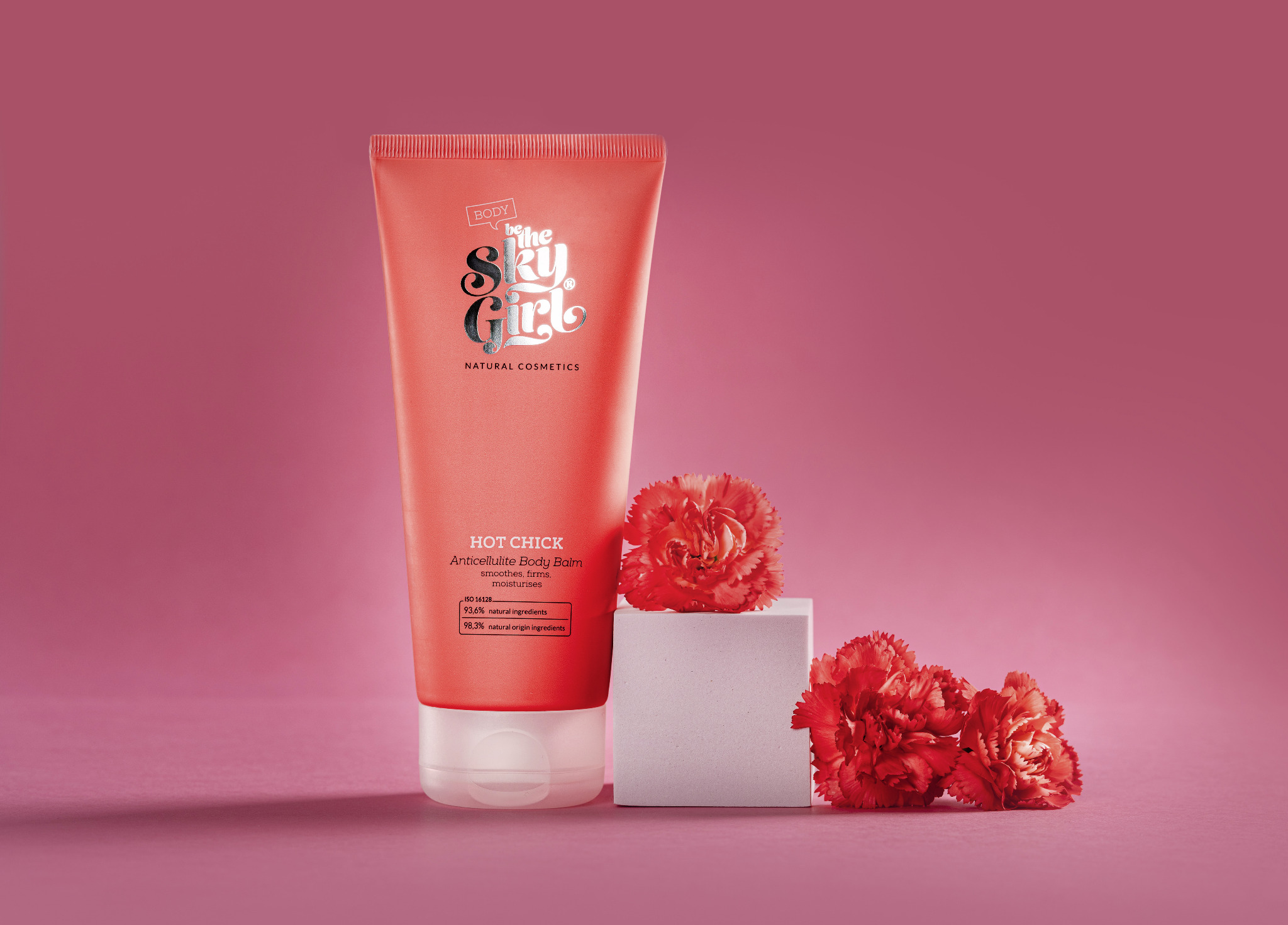 Zdjęcie tubki balsamu antycellulitowego 'The Sky Girl' z Marki, umieszczonej obok dwóch czerwonych goździków i białego sześcianu, na jednolitym różowym tle.