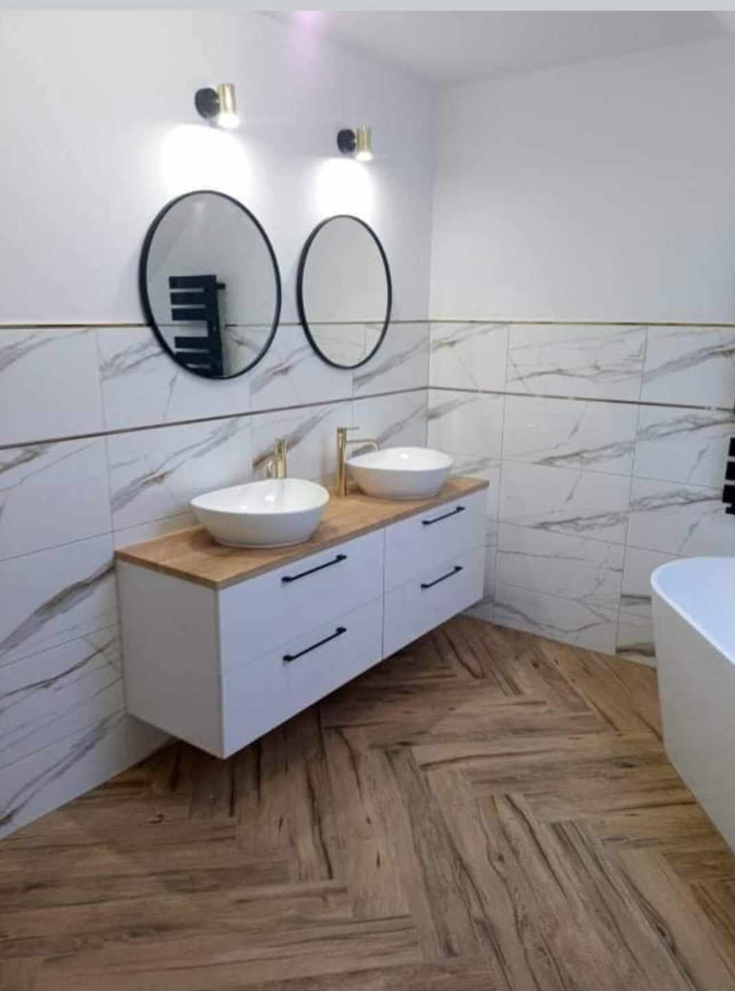 Nowoczesna łazienka z dwiema okrągłymi umywalkami nablatowymi, złotą armaturą i białymi szafkami z czarnymi uchwytami, wykończona płytkami imitującymi marmur z cienką złotą linią oraz parkietem...