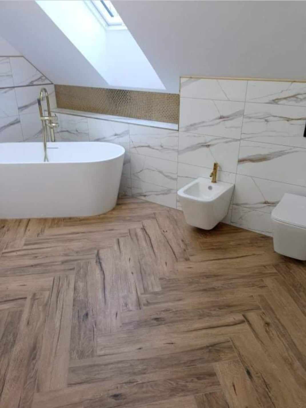 Nowoczesna łazienka na poddaszu z drewnopodobną podłogą ułożoną w jodełkę, białą wanną wolnostojącą i marmurowymi płytkami ściennymi z złotymi akcentami.